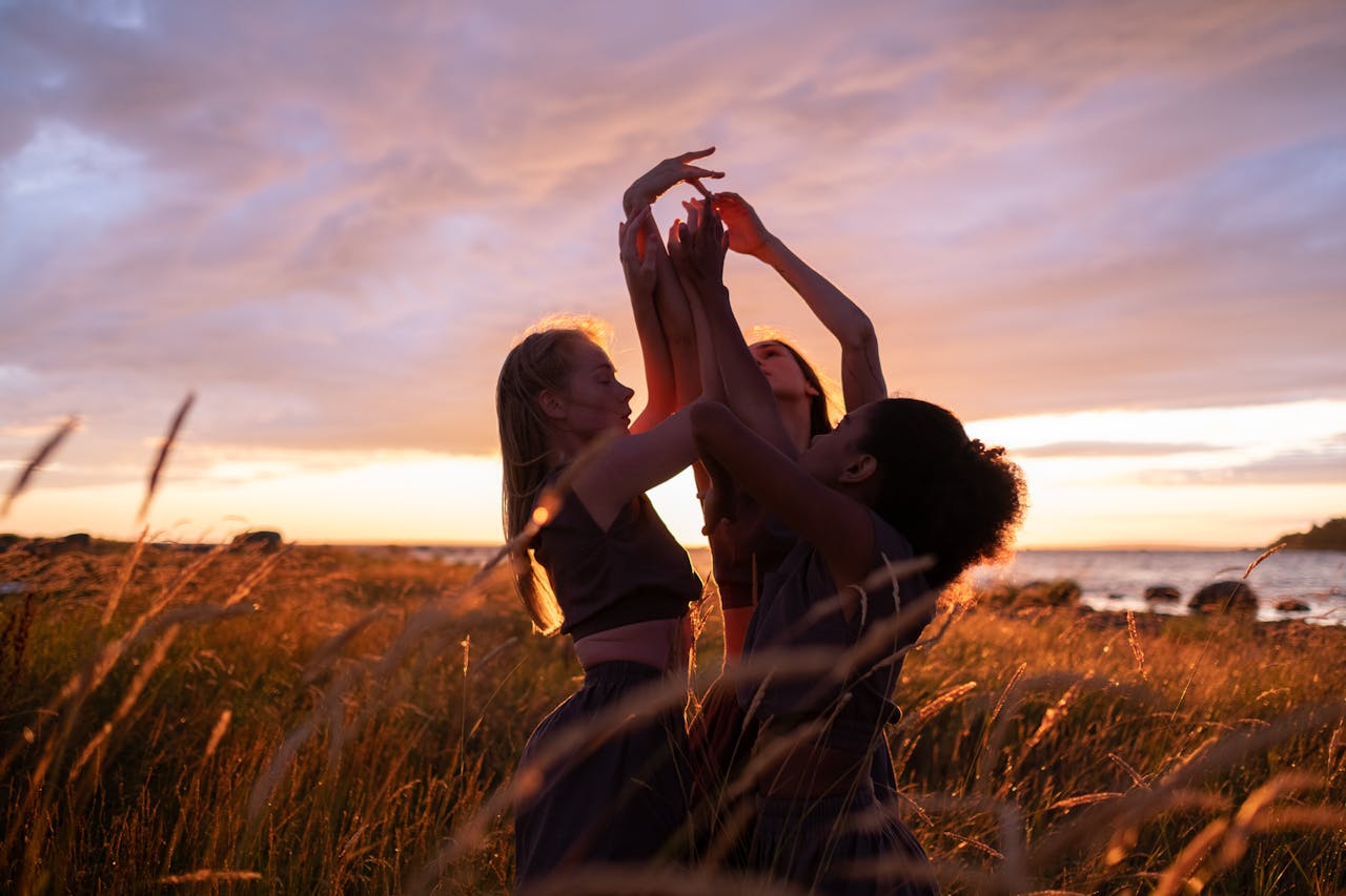 Three women gracefully dancing in a rural meadow as the sun sets behind them.
