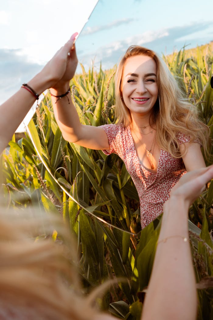 Crop cheerful stylish female with mirror standing among colorful plants in field and looking at camera