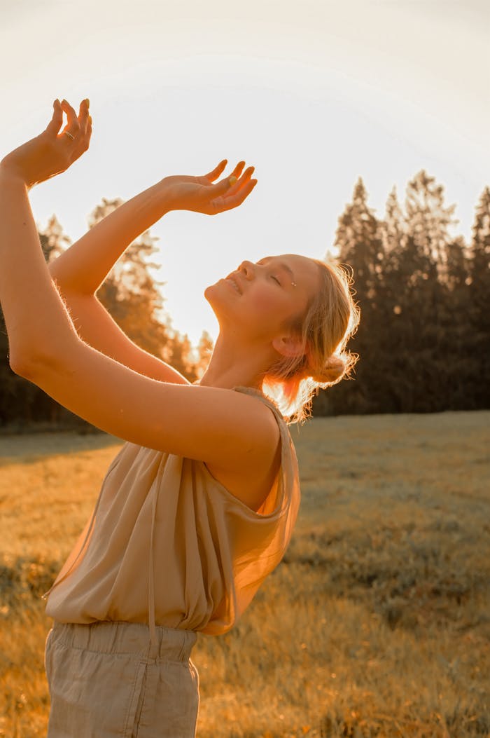 A serene woman basks in the warm glow of sunset with arms raised, standing in a grassy field.